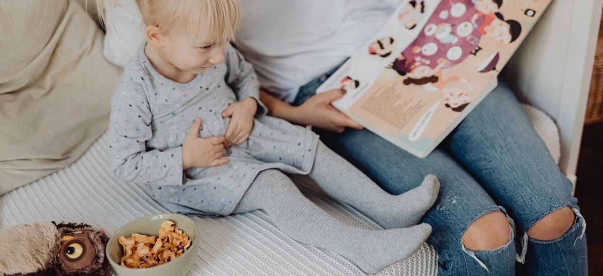 Mother reading a book to her daughter in a cozy bedroom setting with snacks nearby.